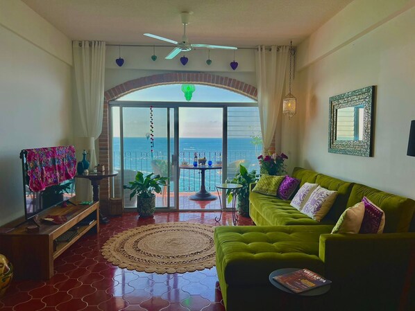 Un salon avec un canapé vert, une table basse en bois et une vue sur la mer, visible à travers une porte vitrée.