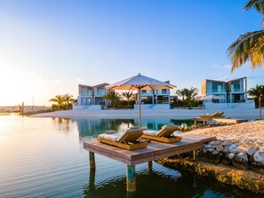A modern resort with a pool, lounge chairs, and a wooden deck over water. - Lagoon Villa 5 Bedroom Including Guesthouse LV16 (Providenciales)