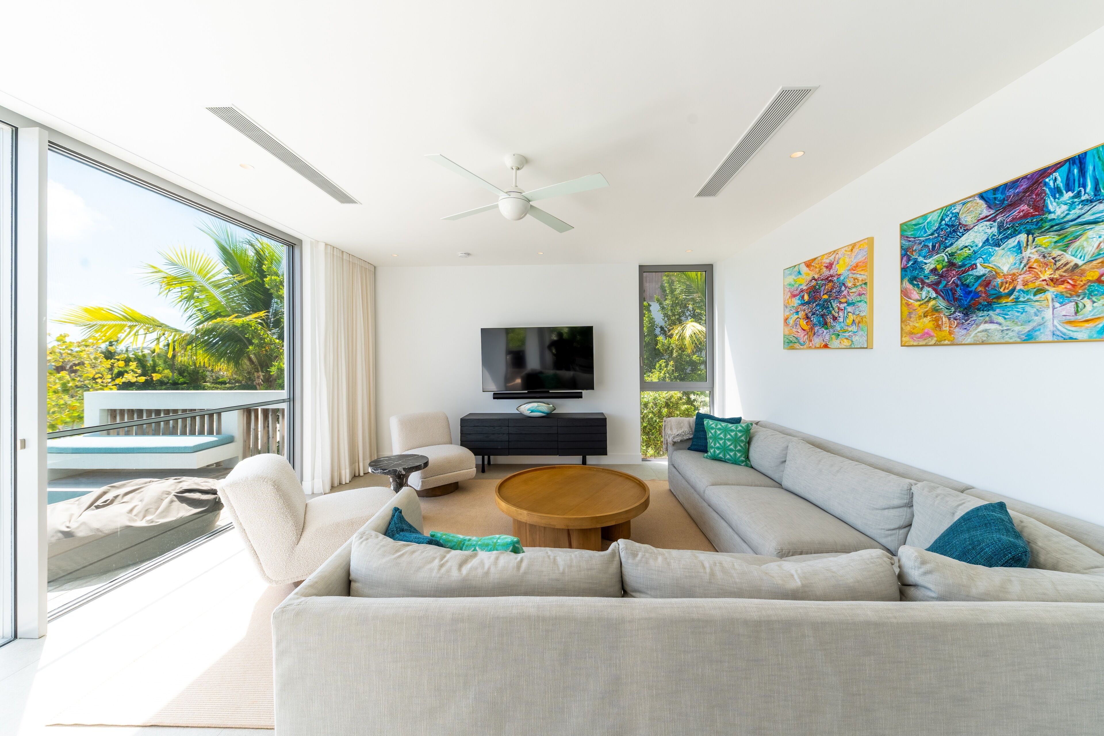 A modern living room with a large sofa, a coffee table, and a flat-screen TV. There is a balcony with a view of palm trees.