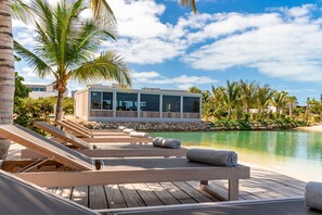 A wooden deck with lounge chairs and a poolside view of a modern building with large windows. - Lagoon Villa 5 Bedroom Including Guesthouse LV16 (Providenciales)