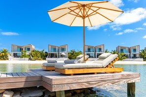 A wooden deck with lounge chairs and an umbrella, overlooking a pool and modern buildings. - Lagoon Villa 5 Bedroom Including Guesthouse LV16 (Providenciales)