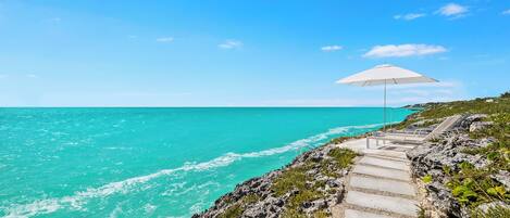 A coastal path leading to a beach with a white umbrella and lounge chairs.