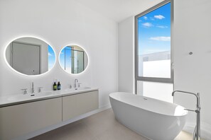 Un baño moderno con una bañera independiente, un tocador doble con dos lavamanos y dos espejos redondos con iluminación integrada.
