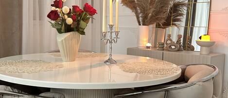 A round dining table with a white lace tablecloth, surrounded by tufted grey chairs.