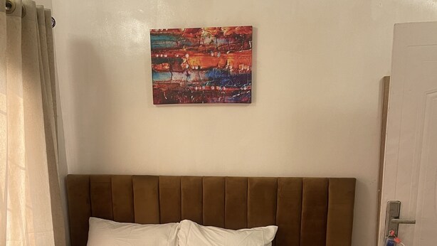 Un lit bien fait, avec du linge de lit blanc, une tête de lit marron et un tableau abstrait aux couleurs vives accroché au mur.