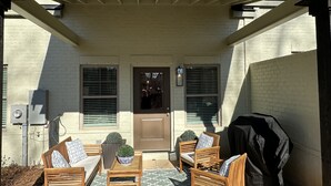 A covered patio area with a brick wall, a door, and two windows with shutters. There are wooden chairs and a small table with a potted plant.