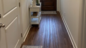 A hallway with a wooden floor, a door with a window, a small table with a lamp, and a rug.