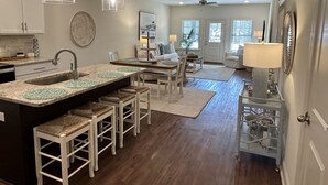 A modern kitchen with a central island, bar stools, pendant lights, and a dining area with a sofa and coffee table.