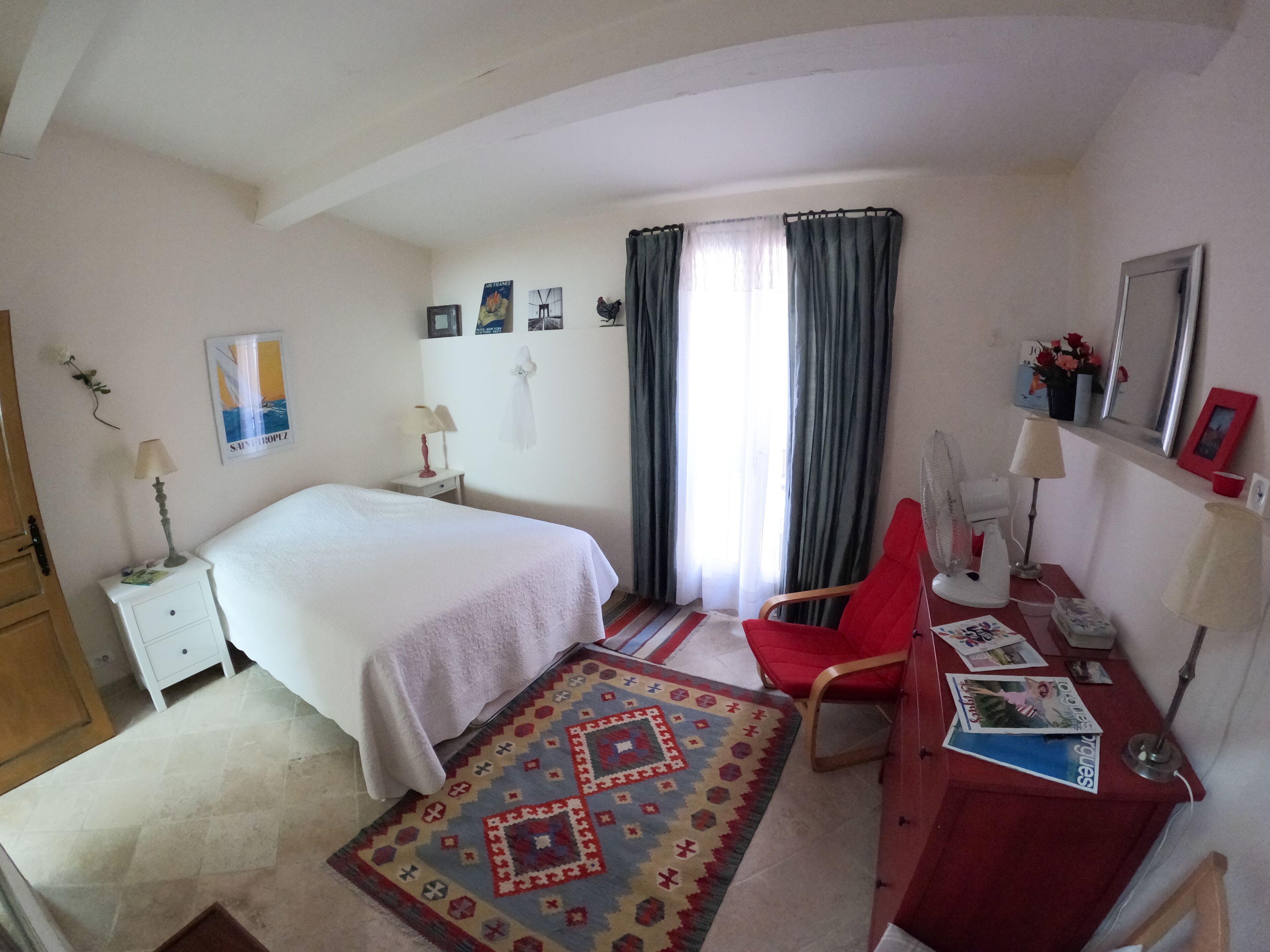 A hotel room with a bed, a desk with a chair, a desk lamp, a red chair, a rug, and a window with curtains.