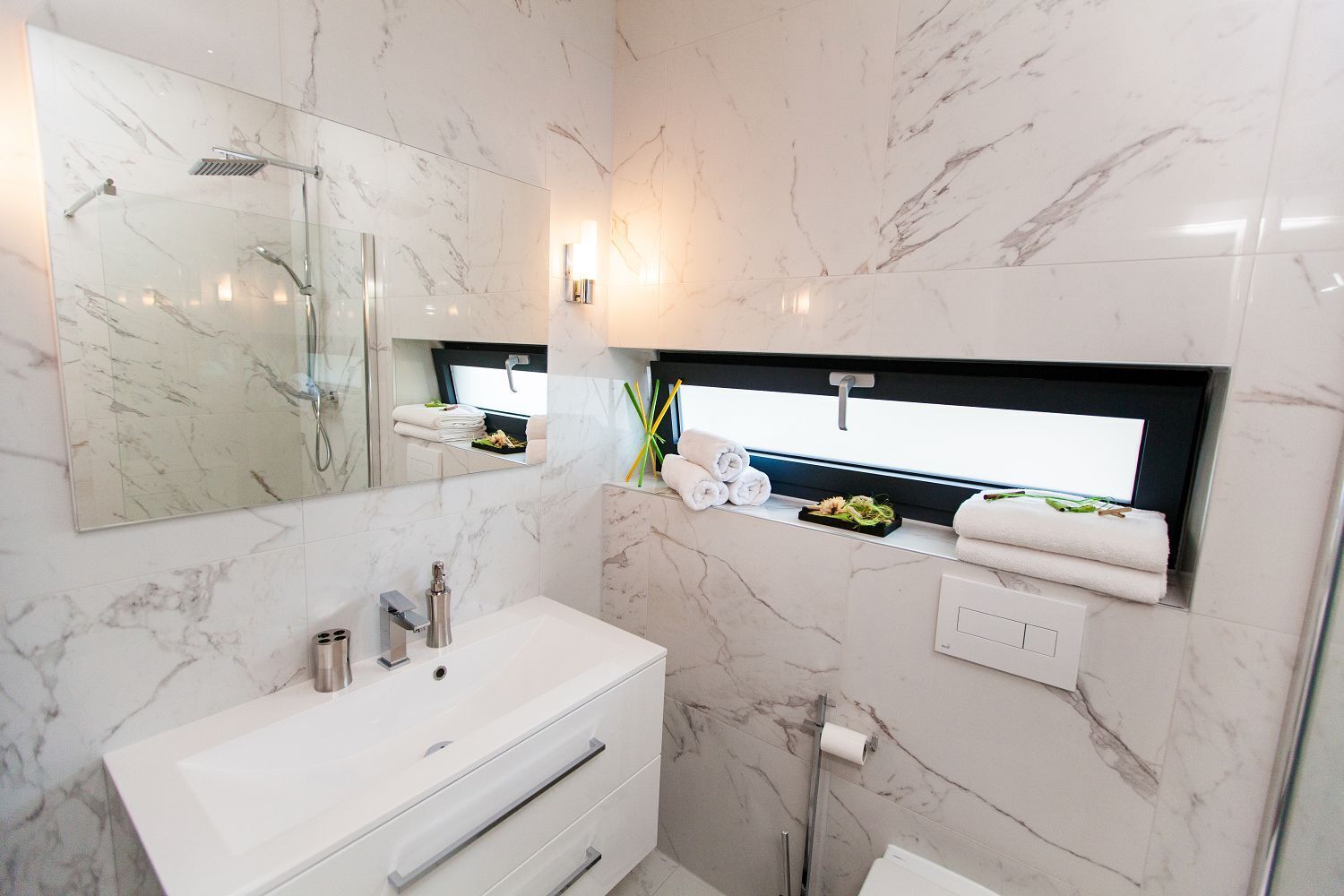 Un baño moderno con tocador de mármol blanco, ducha con cerramiento de vidrio y estante empotrado con toallas y artículos de aseo.