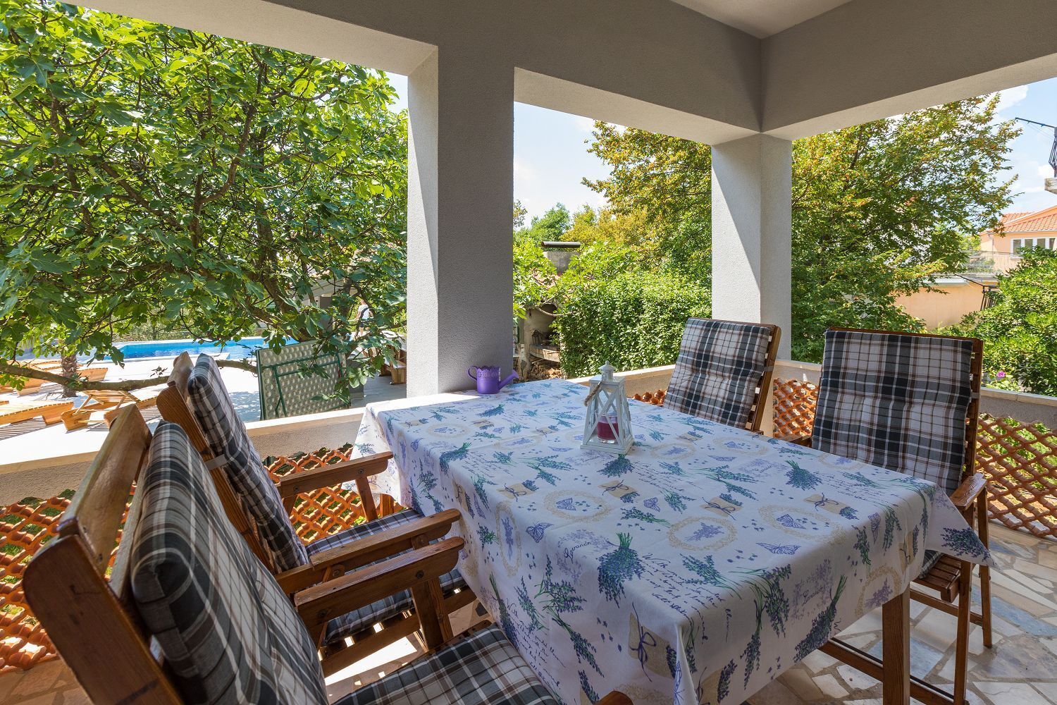 A covered outdoor patio with a table and chairs, surrounded by greenery and a pool.