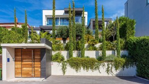 A modern residential building with a wooden gate, a balcony, and a well-maintained garden with tall trees and shrubs.