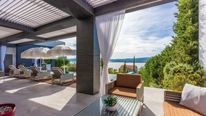 An outdoor lounge area with sun loungers, a table with a potted plant, and a view of the sea and mountains.
