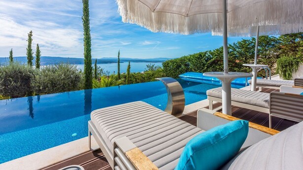 A luxurious infinity pool with a clear view of the sea and mountains, surrounded by modern lounge chairs and a large umbrella.