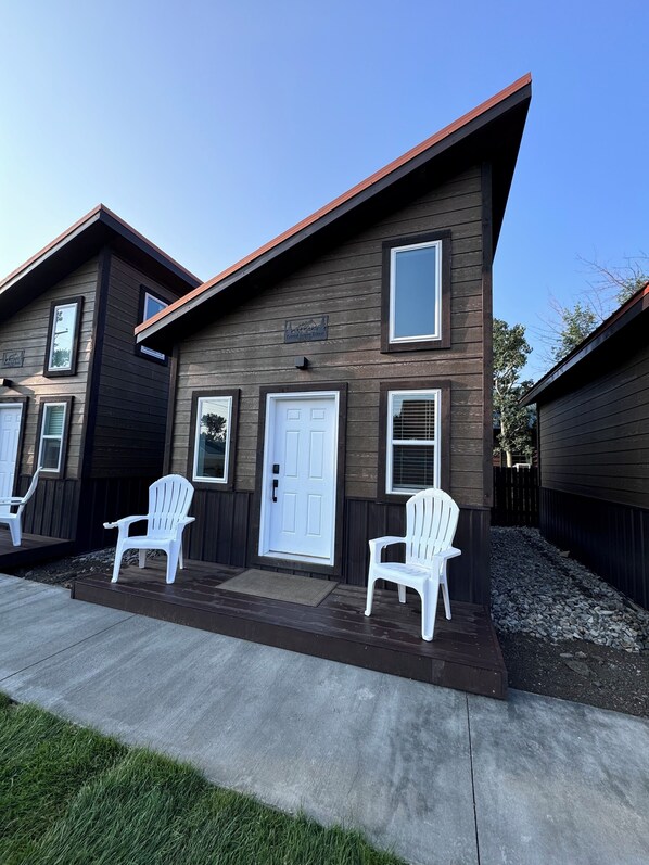 Un bâtiment à deux étages, à l’extérieur de couleur brun foncé, avec une porte d’entrée blanche et deux chaises Adirondack blanches sur le porche avant.