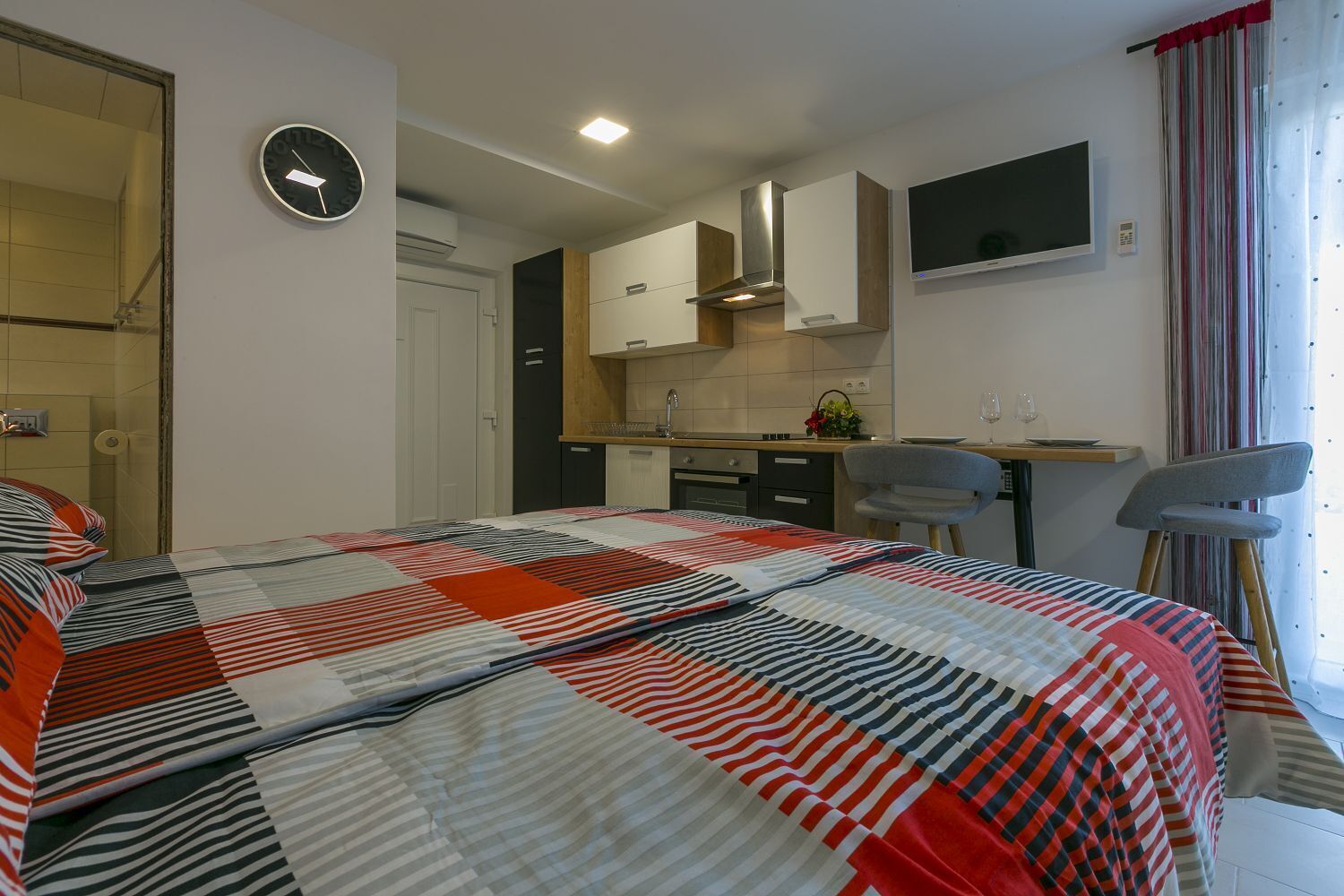 A bedroom with a large bed, a small kitchenette, a TV, and a window with curtains.