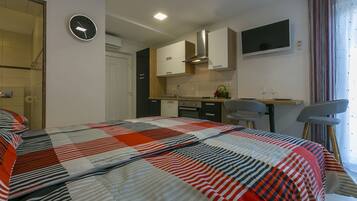 A bedroom with a large bed, a small kitchenette, a TV, and a window with curtains.