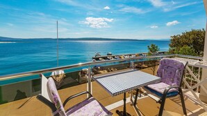 Un balcon avec une table et des chaises donnant sur une marina, où l’on peut admirer des bateaux et une mer d’un bleu limpide.