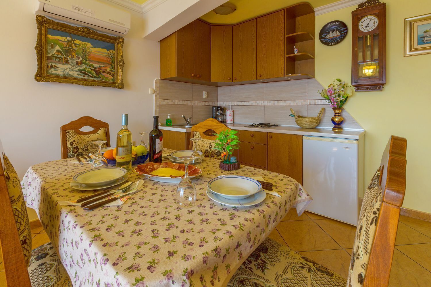 Un comedor con una mesa dispuesta para una comida, armarios de madera, un reloj y una pequeña planta en la encimera.