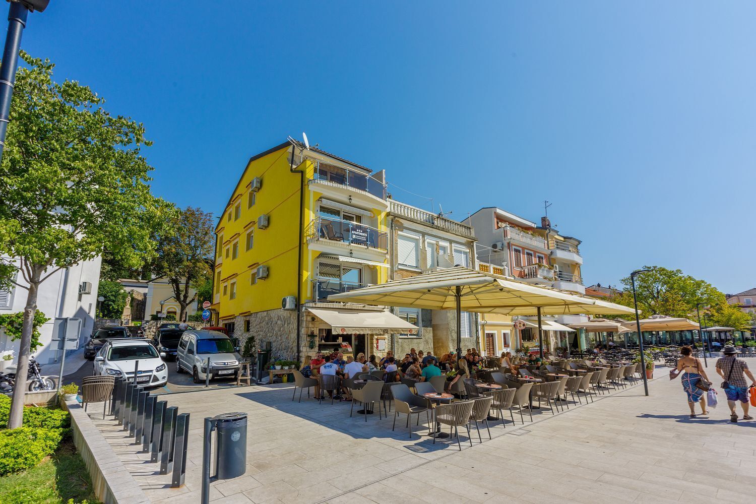 Una plaza pública con mesas al aire libre, un edificio amarillo y un cielo azul despejado.