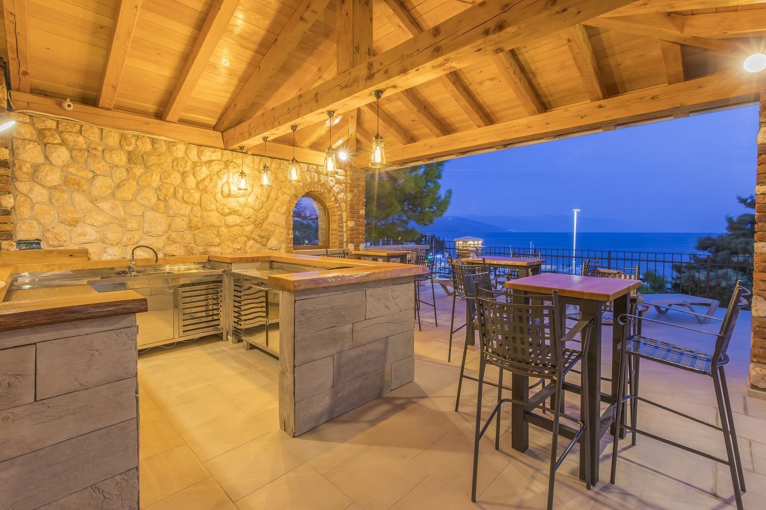 An outdoor kitchen area with stone walls, wooden beams, and a view of the sea.