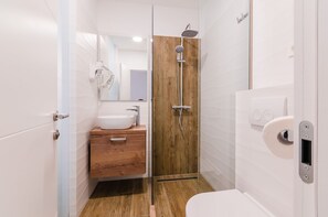 Une salle de bain moderne avec un meuble-lavabo en bois, un grand miroir, un lavabo blanc, une douche équipée d’un pommeau de douche à main et des toilettes.