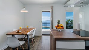 A modern kitchen with a wooden island, white chairs, and a dining table. A fruit bowl is on the island, and a glass of orange juice is on the table.