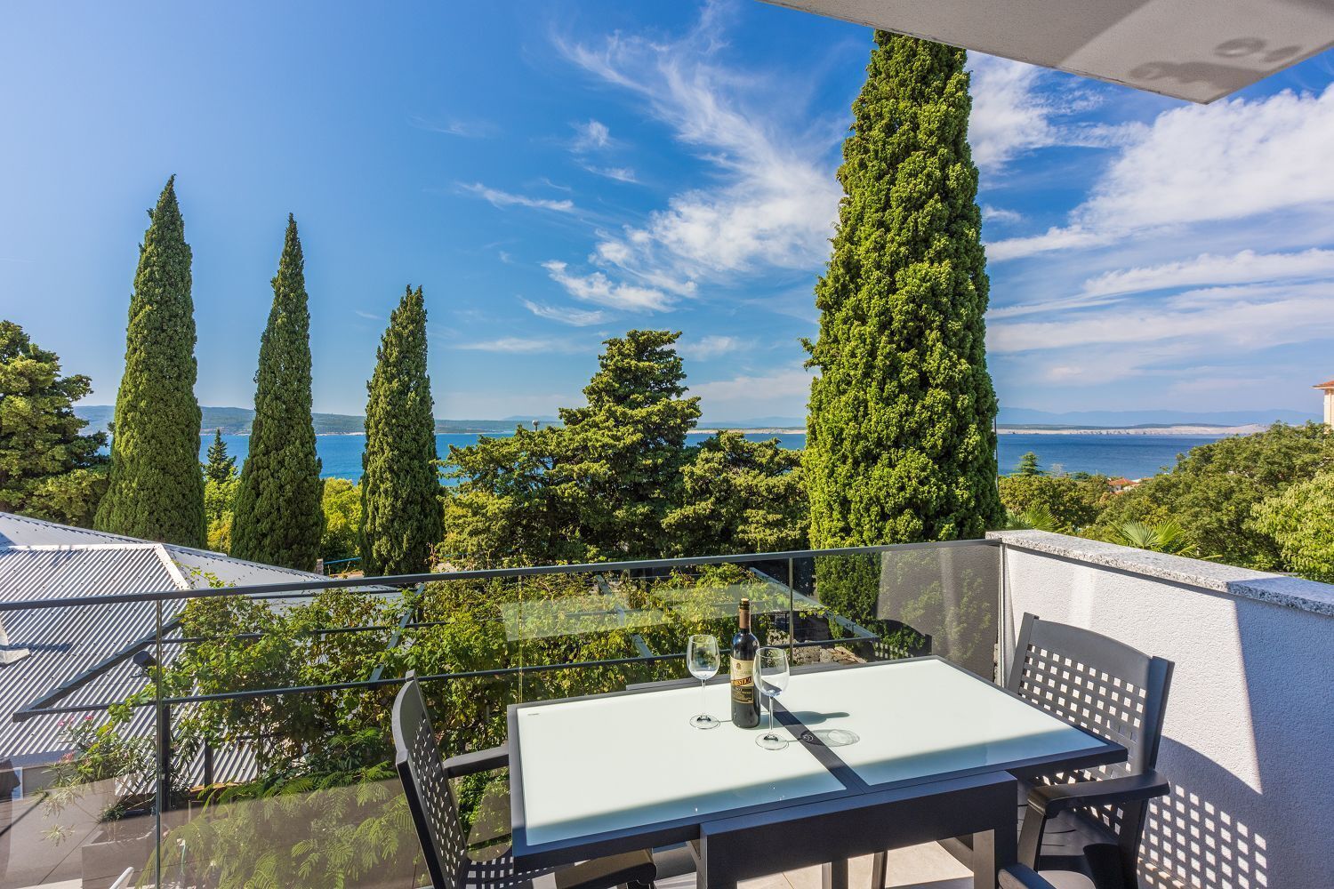 En balkon med et bord og stole, der giver udsigt over et smukt landskab med hav og høje træer.