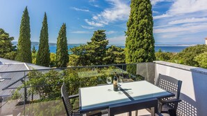 Uma varanda com mesa e cadeiras, com vista para o mar e árvores altas.