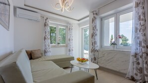 Una sala de estar moderna con un sofá beige, una mesita redonda con fruta y vistas a zonas verdes por las ventanas.