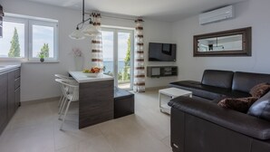 A modern living room with a dark leather sofa, a white dining table with a bowl of fruit, a TV mounted on the wall, and a balcony with curtains. - 500 metres to the beach with pool, sea view (Crikvenica)