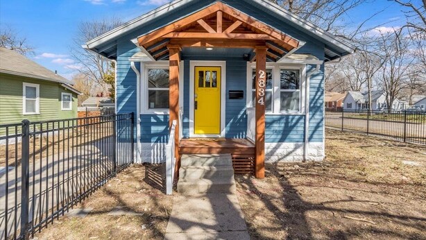 A small, single-story house with a yellow front door, a porch with wooden beams, and a black metal fence surrounding the property. - Walk to Wesley! Pet Friendly + Fully Fenced Lot (Wichita)