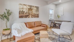 A modern living room with a brown leather sofa, a white armchair, and a large cityscape painting on the wall. - Walk to Wesley! Pet Friendly + Fully Fenced Lot (Wichita)