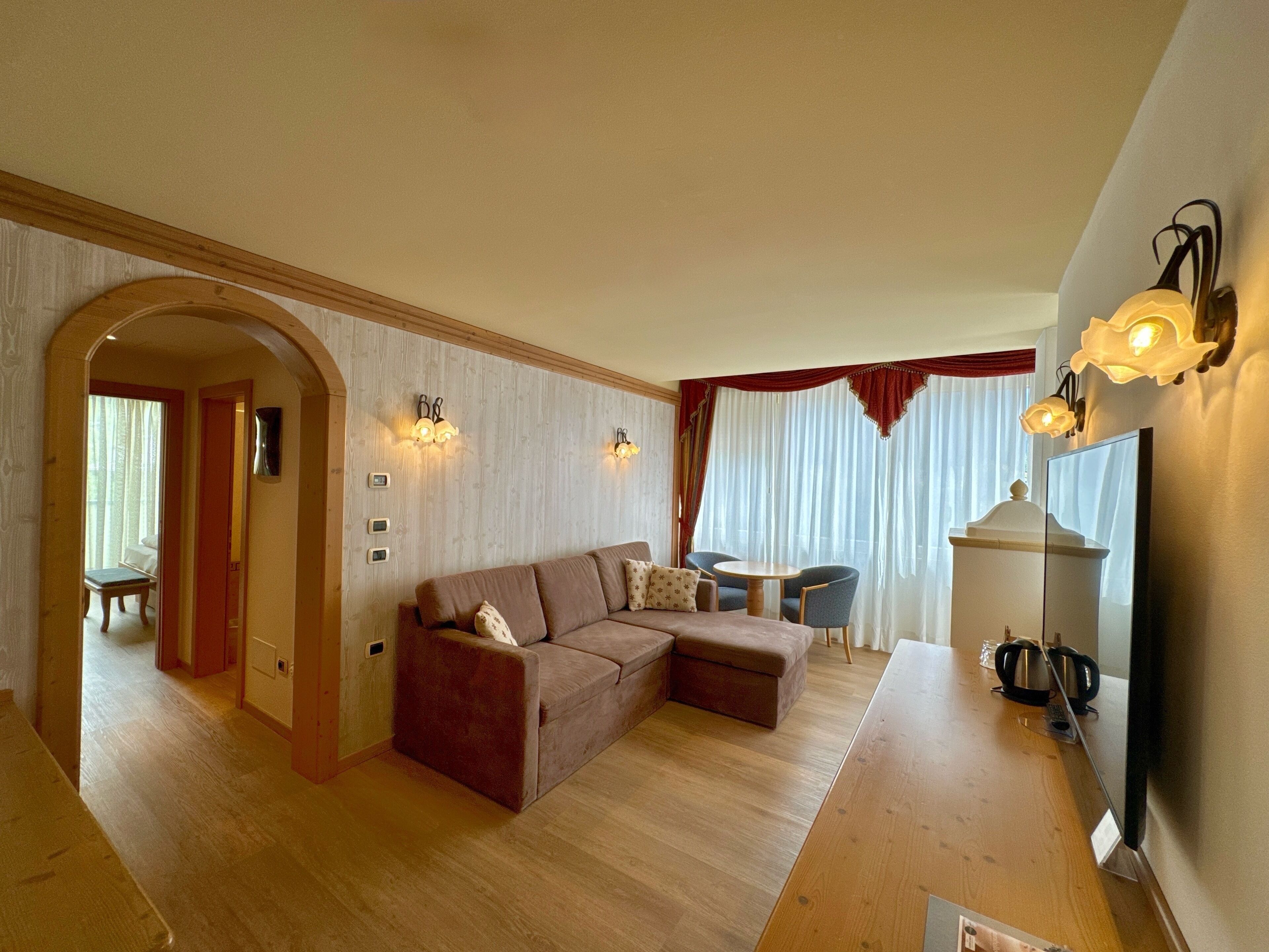 A cozy living room with a brown sectional sofa, a wooden coffee table, and a TV on a wooden stand. There is a dining area with a small table and chairs, and a hallway leading to another room.