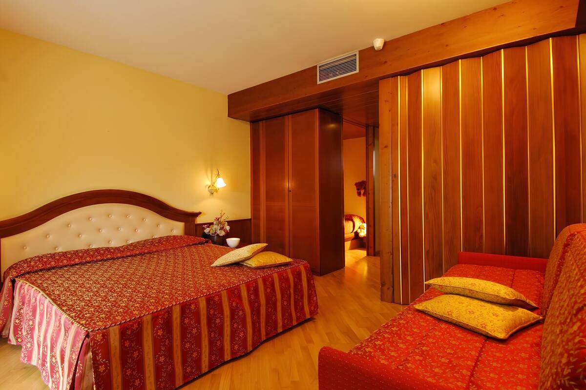 A hotel room with a large bed, a sofa, a small table with a vase, and a wall-mounted lamp.