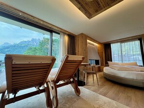 A modern hotel room with a large window, wooden furniture, a sofa, and a fireplace. - Hotel Mirabello - Slow Hotel Benessere (Fiera di Primiero)