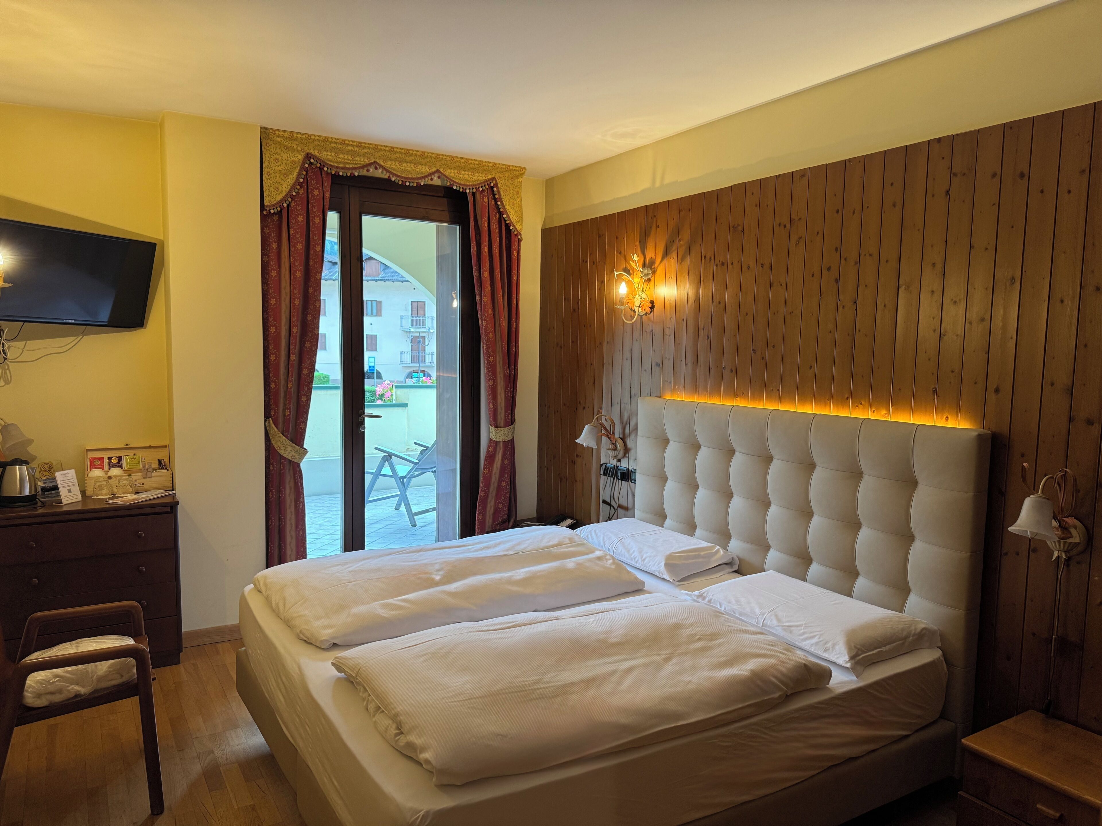 Habitación de hotel con cama, televisor, escritorio con lámpara y vistas al exterior.