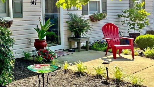 A small, single-story house with a number 147 on the door, a red chair, and various potted plants. - Parkers Flat 147, Comfy and Cozy.  Fully Furnished , Sparkling Clean . (Aiken)
