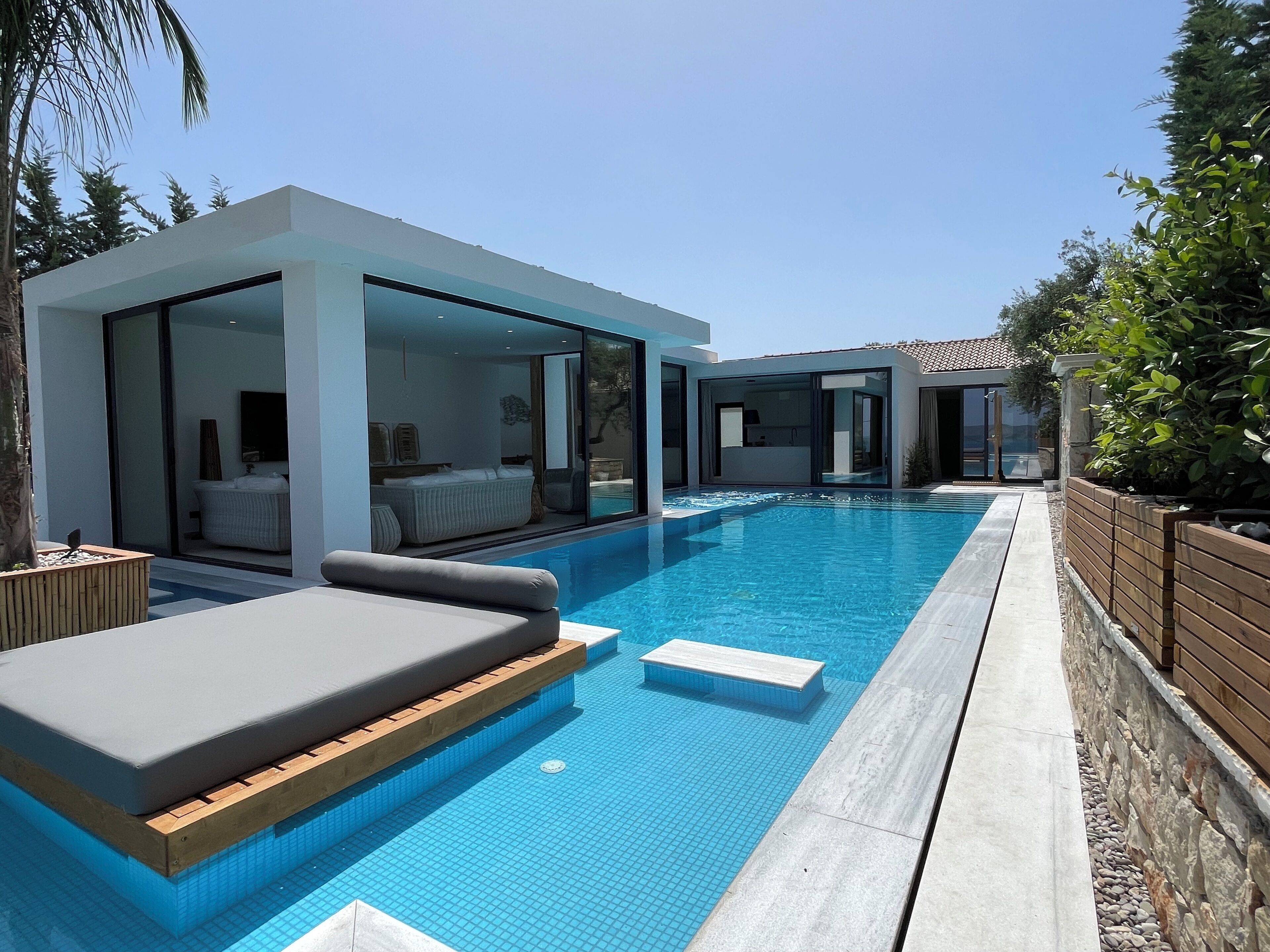 Un espace moderne aménagé au bord de la piscine, avec une piscine à l’eau d’un bleu limpide, une terrasse en bois et une chambre fermée par des parois de verre, où l’on peut voir un lit et une salle de bain.