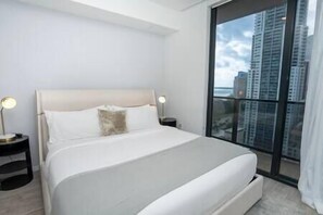 A modern bedroom with a large bed, two bedside tables, and a view of tall buildings through a large window.