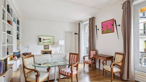 A well-lit living room with a glass-top coffee table, striped chairs, a white sofa, and a bookshelf filled with books. - Chez Patrick (Paris)