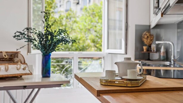 A modern kitchen with a view of buildings through a large window, a wooden table with a tray of white cups and a teapot, and a vase with green foliage. - Chez Patrick (Paris)