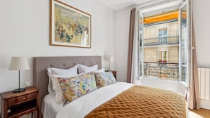 A hotel room with a bed, a nightstand, a lamp, a window with a view of a building, and a painting on the wall. - Chez Patrick (Paris)