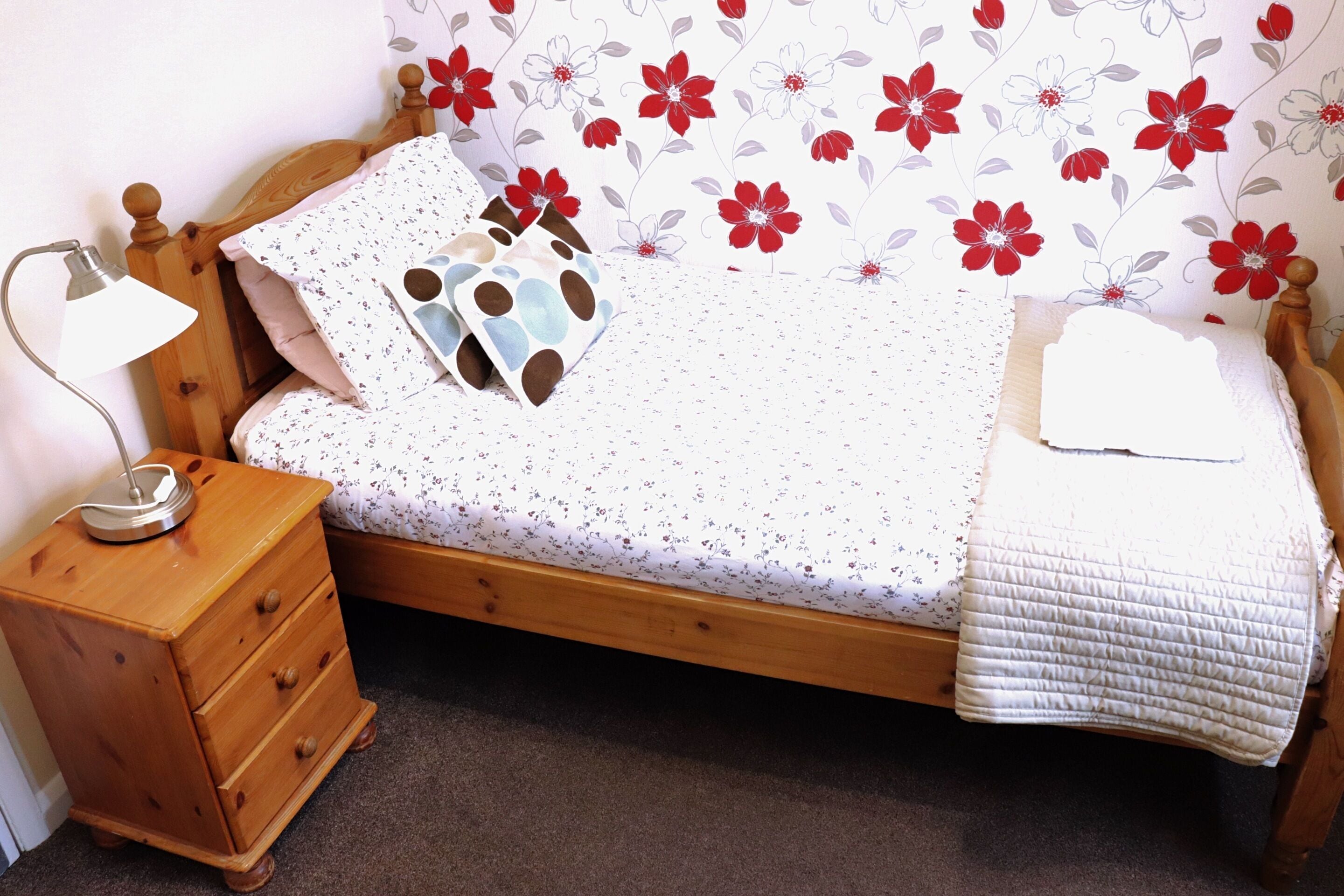 Een keurig opgemaakt bed met een hoofdbord met bloemenprint, een nachtkastje met lades en een enkele bedlamp.