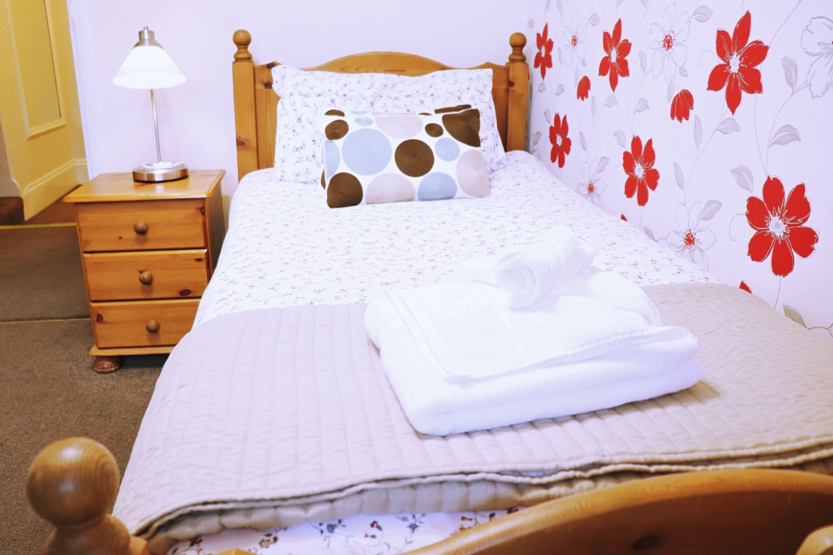 A neatly made bed with a floral-patterned duvet, a bedside lamp, and a wooden nightstand with drawers.
