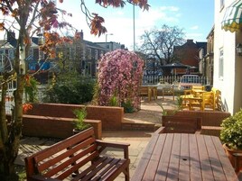 An outdoor patio area with wooden benches and tables, surrounded by brick planters and flowering plants.