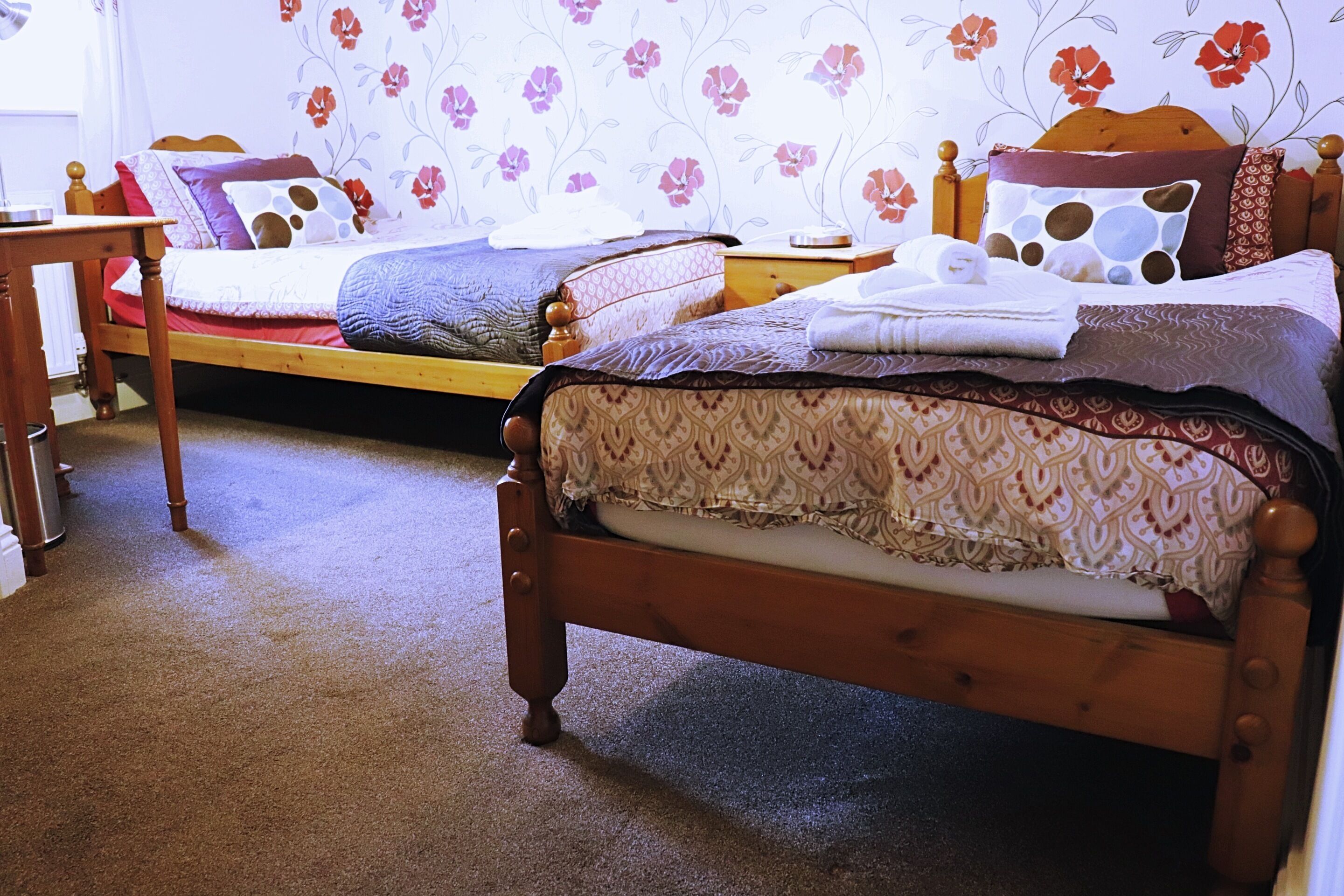 Ein Zimmer mit zwei Holzbett, jedes mit gemusterten Bettbezügen und Kissen. Es gibt einen Nachttisch aus Holz und einen Tisch mit einer Lampe.
