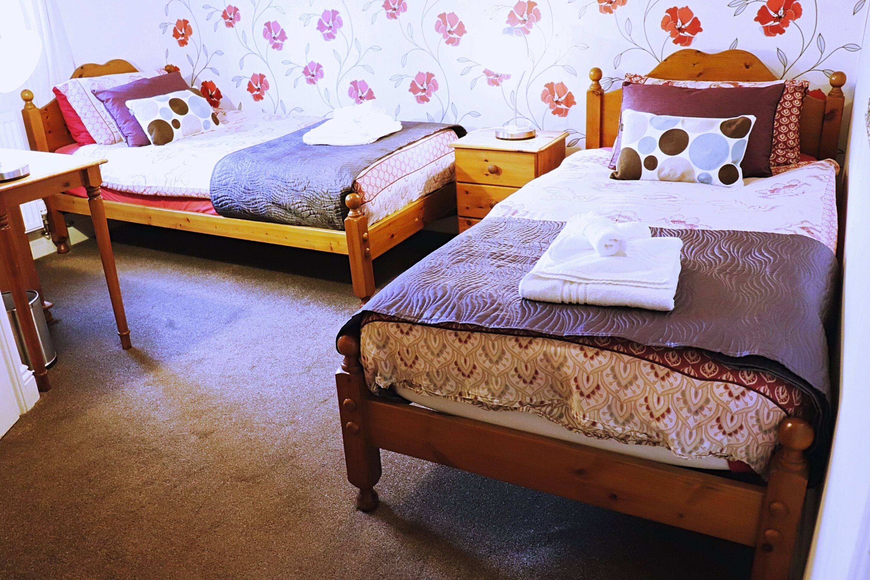 A bedroom with two wooden beds, each with a patterned bedspread and pillows. There is a wooden nightstand with a lamp and a small table with a lamp. The room has floral wallpaper and a carpeted floor.