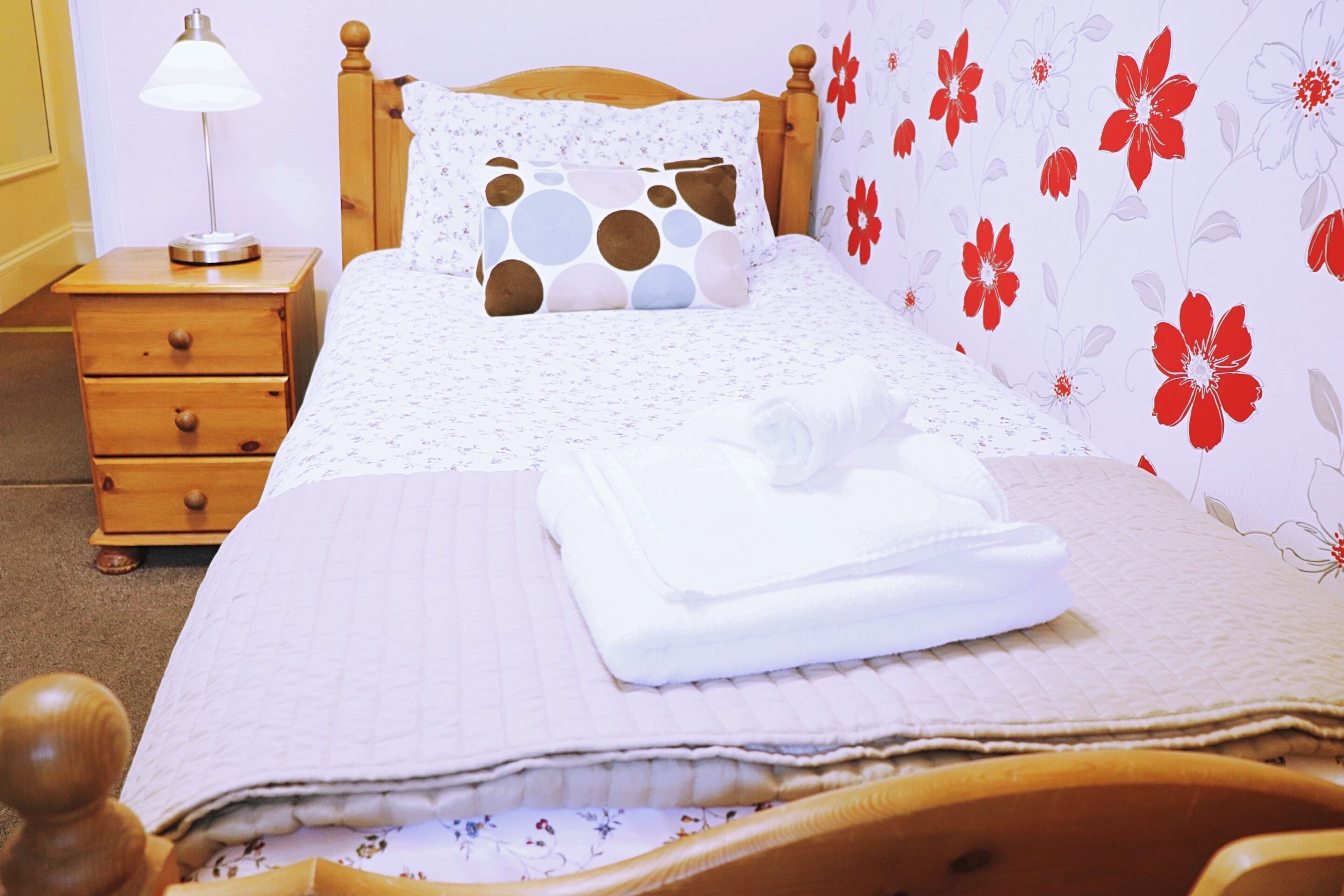 Una cama bien tendida con un papel tapiz estampado con motivos florales y una mesita de noche de madera con una lámpara.