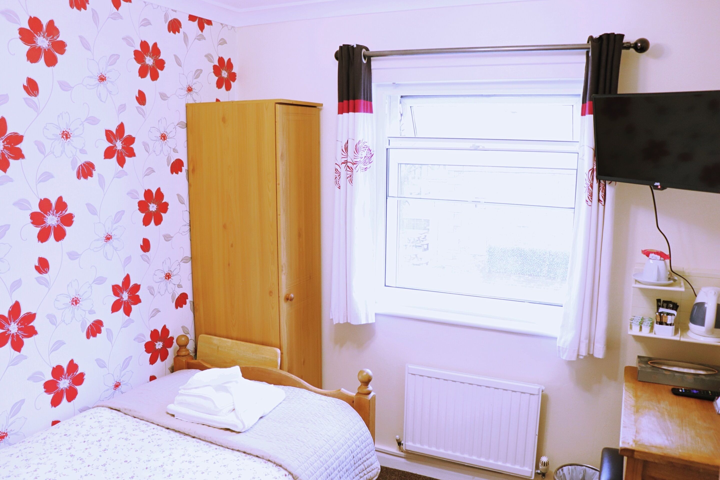 Une chambre à coucher comprenant un lit, une armoire, une fenêtre avec des rideaux, un radiateur et une étagère avec divers objets.